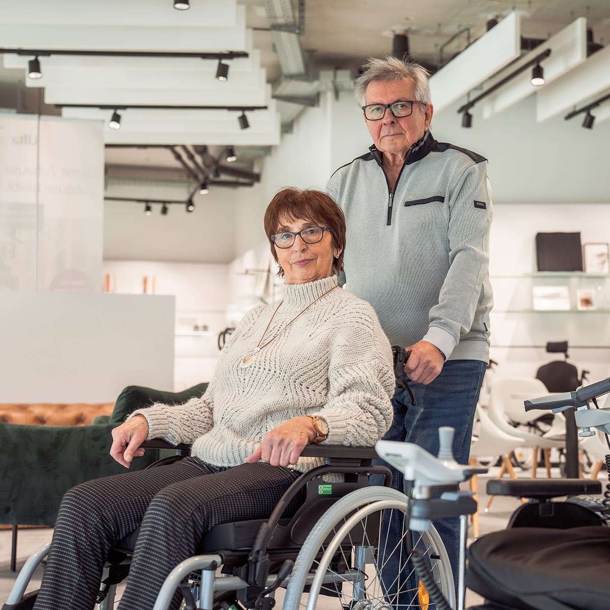 Ältere Frau im Rollstuhl, dahinter steht ein älterer Mann, in der Ausstellung